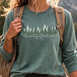 Può includere: Una felpa a maniche lunghe girocollo color verde salvia con una grafica raffigurante la silhouette di una foresta in tonalità di bianco e verde scuro. Sotto gli alberi, il testo recita YELLOWSTONE NATIONAL PARK EST. 1872 in un font bianco sans-serif.