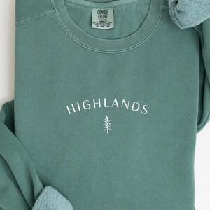May include: A teal green sweatshirt featuring the word "HIGHLANDS" in white script and a small tree graphic. The sweatshirt has a crew neck and long sleeves. A tag on the inside collar reads "COMFORT COLORS".