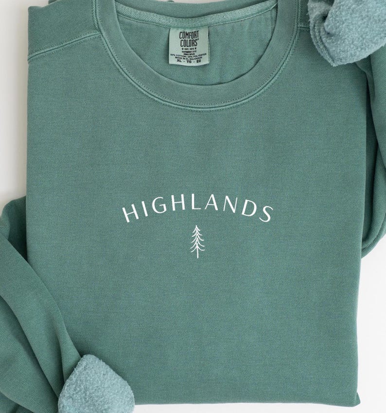 May include: A teal green sweatshirt featuring the word "HIGHLANDS" in white script and a small tree graphic. The sweatshirt has a crew neck and long sleeves. A tag on the inside collar reads "COMFORT COLORS".