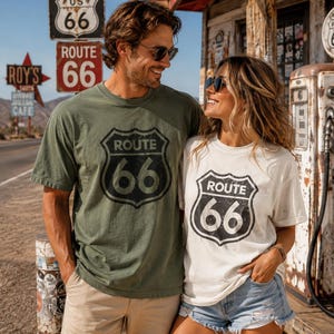Puede incluir: Una camiseta verde oliva y una camiseta blanca, ambas con un gr&aacute;fico negro desgastado en forma de escudo de la Ruta 66 en el pecho. Las camisetas tienen mangas cortas y cuello redondo, mostrando el dise&ntilde;o ic&oacute;nico de se&ntilde;al de tr&aacute;fico con el texto Route 66.