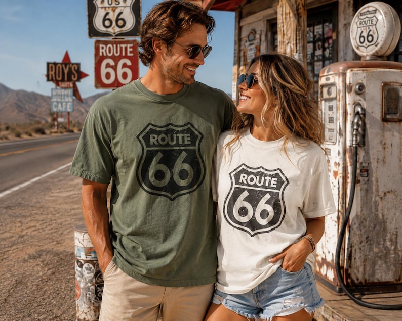Puede incluir: Una camiseta verde oliva y una camiseta blanca, ambas con un gr&aacute;fico negro desgastado en forma de escudo de la Ruta 66 en el pecho. Las camisetas tienen mangas cortas y cuello redondo, mostrando el dise&ntilde;o ic&oacute;nico de se&ntilde;al de tr&aacute;fico con el texto Route 66.