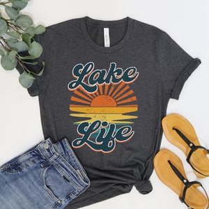 May include: Dark gray t-shirt with the words "Lake Life" in teal script, above a retro sunset graphic in orange, yellow, and teal. The shirt is paired with blue jeans and sandals.
