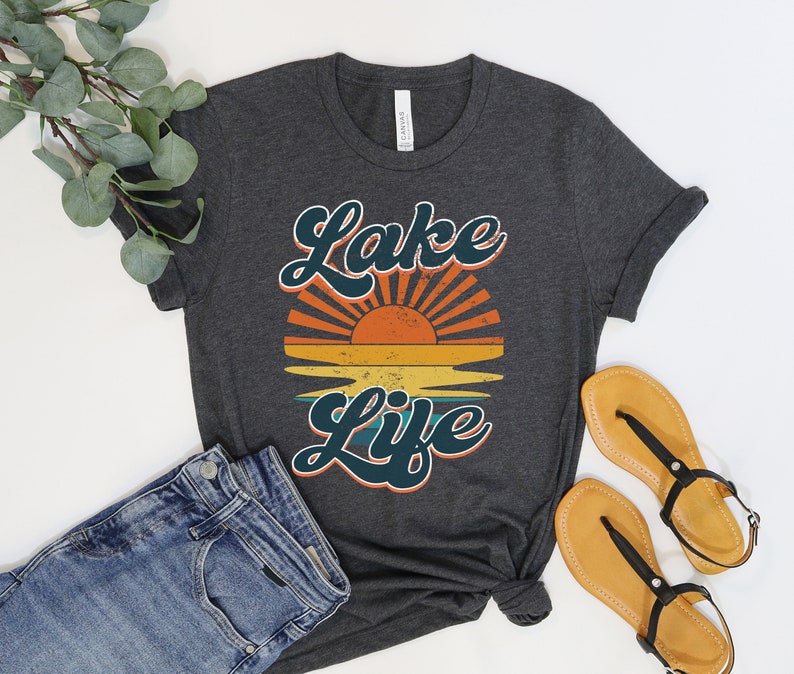 May include: Dark gray t-shirt with the words "Lake Life" in teal script, above a retro sunset graphic in orange, yellow, and teal. The shirt is paired with blue jeans and sandals.