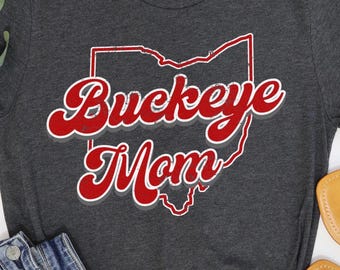 Pode incluir: Uma t-shirt cinzenta escura mesclada com um gráfico desgastado em vermelho e branco do estado de Ohio. O texto "Buckeye Mom" está impresso numa fonte cursiva vermelha e arrojada com uma sombra cinzenta sobre o contorno do estado à frente.