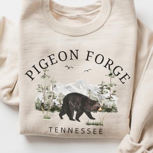 K&ouml;nnte beinhalten: Ein cremefarbenes Sweatshirt mit Rundhalsausschnitt und einer schwarzen Grafik eines Schwarzb&auml;ren, der durch einen Wald mit Bergen im Hintergrund l&auml;uft. Der Text PIGEON FORGE ist &uuml;ber dem B&auml;ren gew&ouml;lbt und TENNESSEE steht darunter.