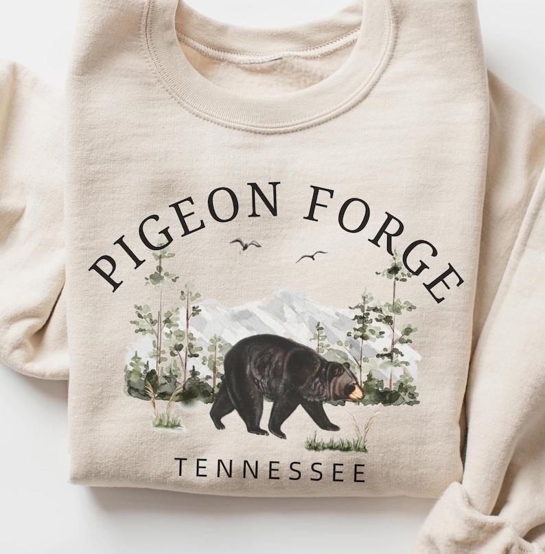 K&ouml;nnte beinhalten: Ein cremefarbenes Sweatshirt mit Rundhalsausschnitt und einer schwarzen Grafik eines Schwarzb&auml;ren, der durch einen Wald mit Bergen im Hintergrund l&auml;uft. Der Text PIGEON FORGE ist &uuml;ber dem B&auml;ren gew&ouml;lbt und TENNESSEE steht darunter.