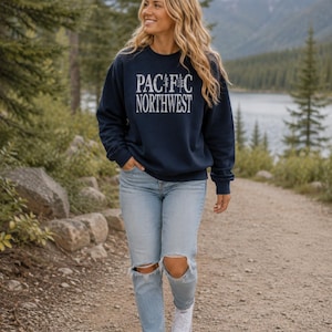 May include: A navy blue crewneck sweatshirt featuring the white text PACIFIC NORTHWEST with a pine tree graphic replacing the letter I, paired with light blue distressed denim jeans and white canvas sneakers.