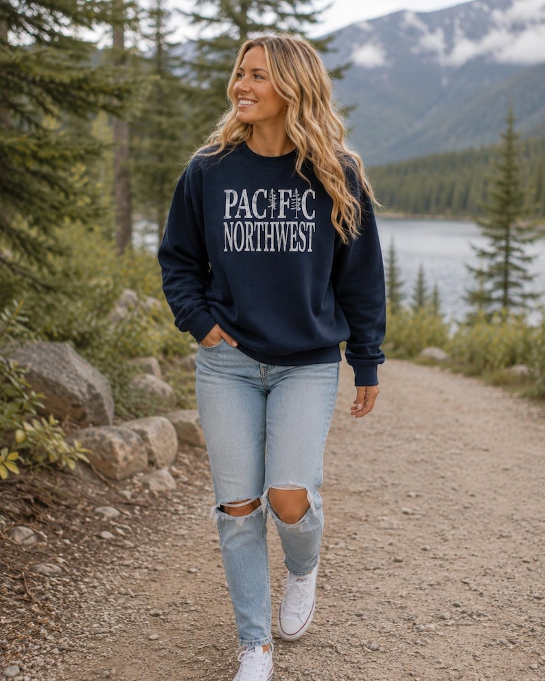 May include: A navy blue crewneck sweatshirt featuring the white text PACIFIC NORTHWEST with a pine tree graphic replacing the letter I, paired with light blue distressed denim jeans and white canvas sneakers.