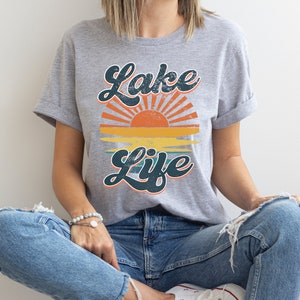 May include: A gray t-shirt with the words "Lake Life" in a retro font, featuring a sunset design with orange, yellow, and teal colors. The shirt is made of a soft material.