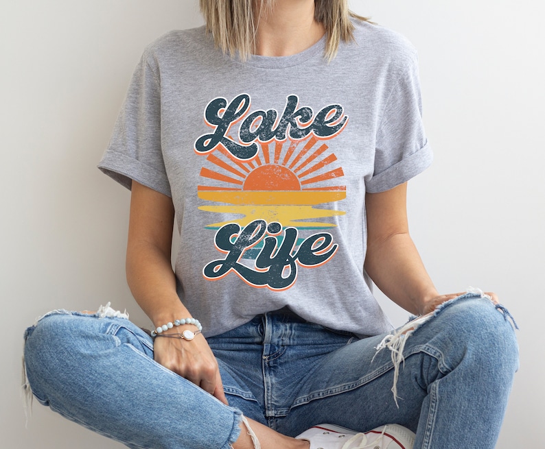 May include: A gray t-shirt with the words "Lake Life" in a retro font, featuring a sunset design with orange, yellow, and teal colors. The shirt is made of a soft material.