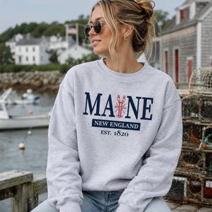 May include: A light grey crewneck sweatshirt featuring dark blue text that reads MAINE with a red lobster graphic replacing the letter I. Below the text is a dark blue rectangle with the words NEW ENGLAND and EST. 1820 printed in white.