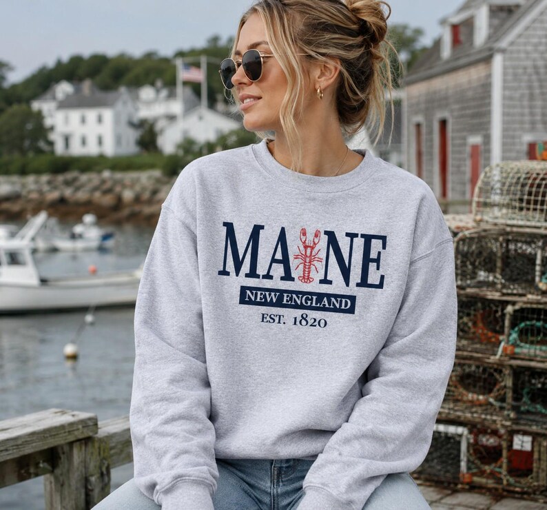May include: A light grey crewneck sweatshirt featuring dark blue text that reads MAINE with a red lobster graphic replacing the letter I. Below the text is a dark blue rectangle with the words NEW ENGLAND and EST. 1820 printed in white.