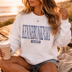 K&ouml;nnte beinhalten: Ein wei&szlig;es Sweatshirt mit dem Text "KENNEBUNKPORT MAINE" in blauen Blockbuchstaben. Das Sweatshirt hat einen Rundhalsausschnitt. Eine beige Kaffeetasse wird in der linken Hand gehalten.