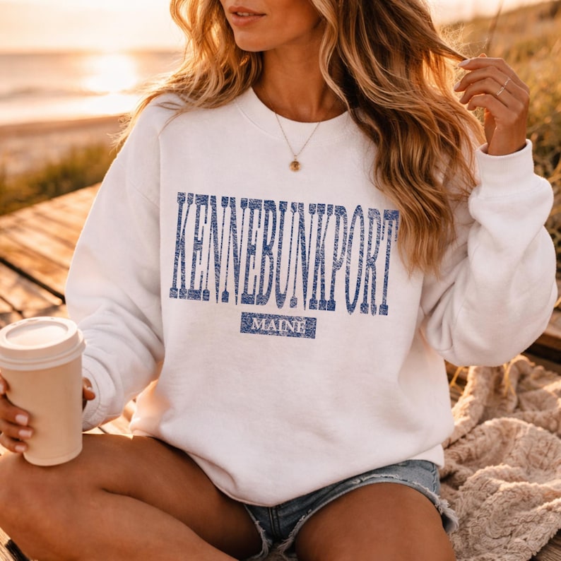 K&ouml;nnte beinhalten: Ein wei&szlig;es Sweatshirt mit dem Text "KENNEBUNKPORT MAINE" in blauen Blockbuchstaben. Das Sweatshirt hat einen Rundhalsausschnitt. Eine beige Kaffeetasse wird in der linken Hand gehalten.