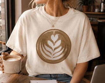 Puede incluir: Una camiseta de manga corta color crema con un gráfico circular grande en el pecho. El gráfico muestra un café latte marrón con arte de espuma blanca en un patrón de corazón y hojas. La camiseta tiene cuello redondo y un corte holgado.