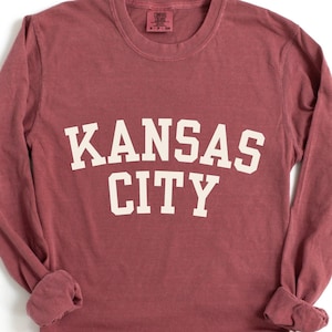 May include: A long-sleeve, maroon-colored shirt with the words "KANSAS CITY" printed in large, white, block letters across the front. The shirt has a crew neck and long sleeves with cuffed wrists.