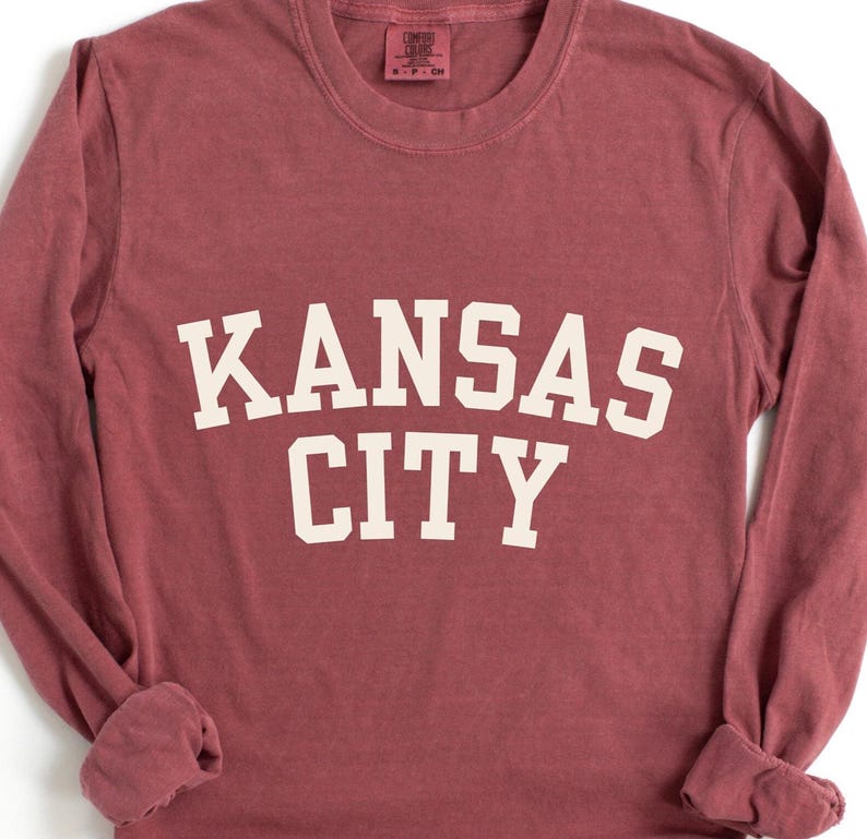 May include: A long-sleeve, maroon-colored shirt with the words "KANSAS CITY" printed in large, white, block letters across the front. The shirt has a crew neck and long sleeves with cuffed wrists.