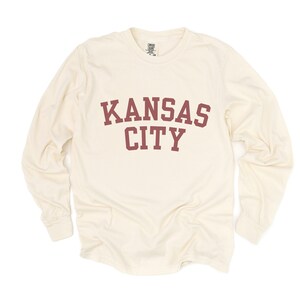 May include: Cream-colored long-sleeve shirt with the words "KANSAS CITY" printed in maroon block letters across the chest. The shirt has a crew neck and long sleeves.