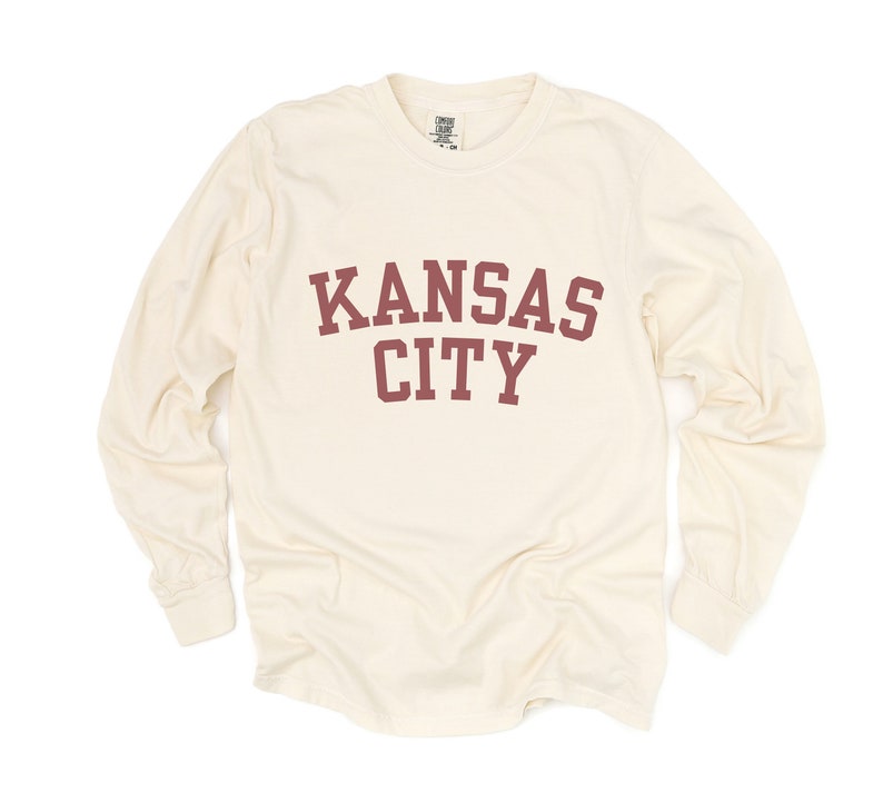 May include: Cream-colored long-sleeve shirt with the words "KANSAS CITY" printed in maroon block letters across the chest. The shirt has a crew neck and long sleeves.
