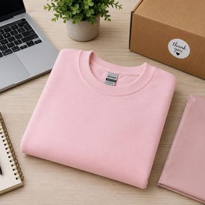 Pode incluir: Uma camisola de gola redonda em rosa claro liso, com mangas compridas e gola canelada. A pe&ccedil;a apresenta uma etiqueta da marca Gildan no interior da gola. A camisola est&aacute; dobrada cuidadosamente e apresentada como uma pe&ccedil;a de vestu&aacute;rio simples.
