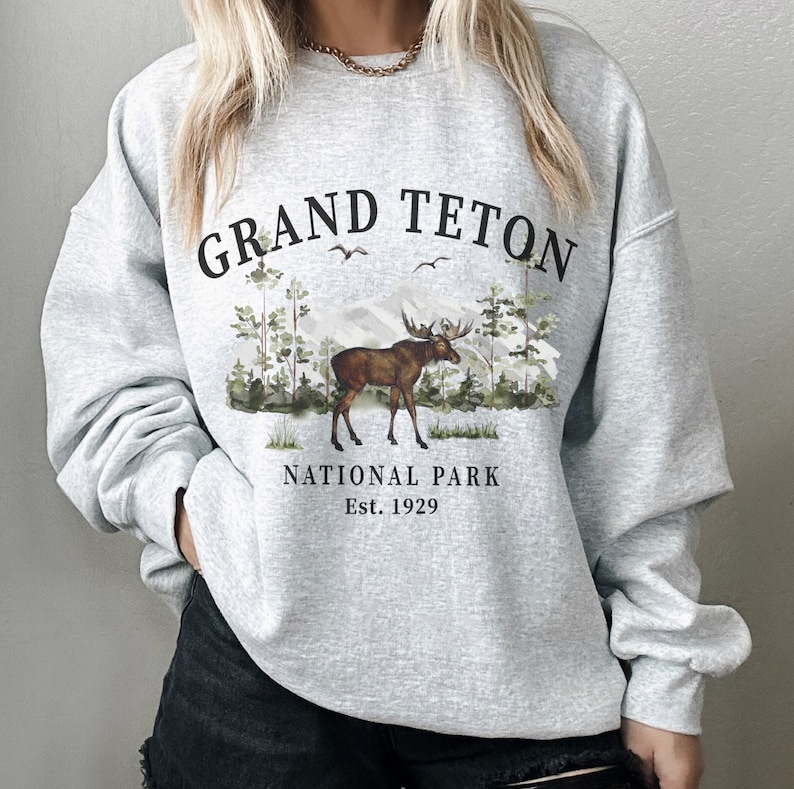 Puede incluir: Sudadera gris claro con la inscripci&oacute;n "GRAND TETON NATIONAL PARK" sobre un gr&aacute;fico de un alce, &aacute;rboles y monta&ntilde;as. La sudadera tambi&eacute;n incluye el texto "Est. 1929".