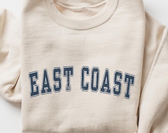 May include: A cream-coloured crewneck sweatshirt featuring the text EAST COAST printed in a dark navy blue collegiate font with a distressed, weathered texture across the chest. The garment has long sleeves and a ribbed collar neckline.