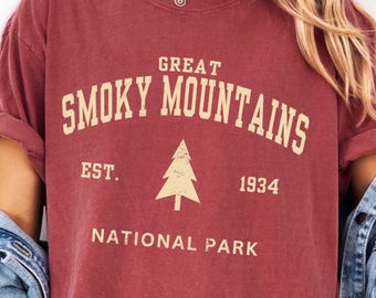 以下が含まれることがあります： ヴィンテージスタイルの「GREAT SMOKY MOUNTAINS NATIONAL PARK」の文字が入ったマルーン色のTシャツ。デザインには木のグラフィックと「1934」の年号が含まれています。シャツは袖をまくり、カジュアルなフィット感です。