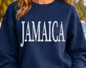 May include: A navy blue crewneck sweatshirt featuring the word JAMAICA printed across the chest in a large, white, distressed serif font. The garment has long sleeves and a ribbed neckline, cuffs, and hem, providing a classic, comfortable fit.