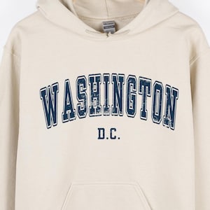 May include: A beige hoodie with the word "WASHINGTON" in navy blue, arched across the chest, with "D.C." below. The hoodie has a drawstring hood and a front pocket.