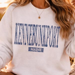 K&ouml;nnte beinhalten: Wei&szlig;es Sweatshirt mit dem Text "KENNEBUNKPORT" in verblichenen blauen Buchstaben. Darunter steht das Wort "MAINE" in einem blauen Rechteck. Das Sweatshirt hat lange &Auml;rmel und einen Rundhalsausschnitt.