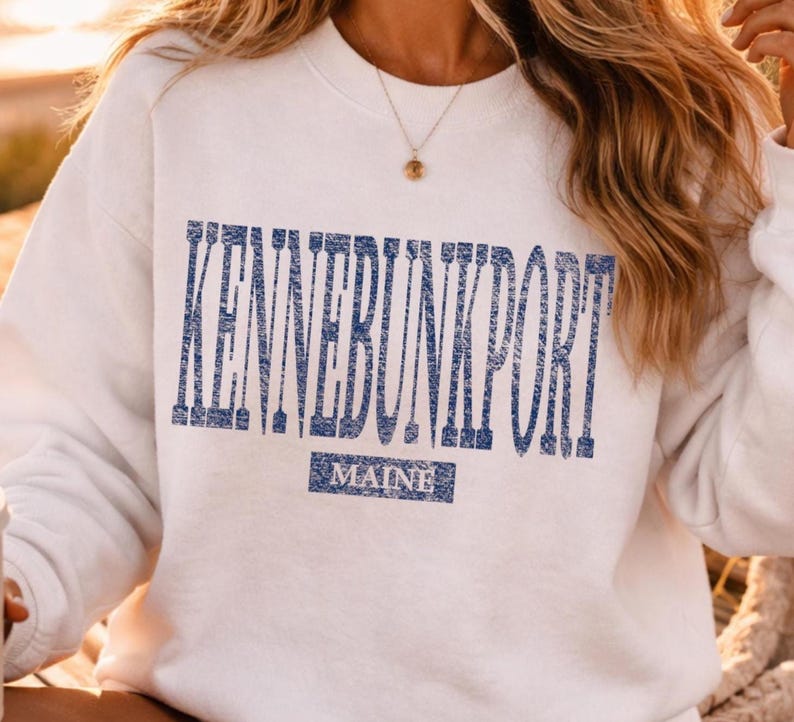K&ouml;nnte beinhalten: Wei&szlig;es Sweatshirt mit dem Text "KENNEBUNKPORT" in verblichenen blauen Buchstaben. Darunter steht das Wort "MAINE" in einem blauen Rechteck. Das Sweatshirt hat lange &Auml;rmel und einen Rundhalsausschnitt.