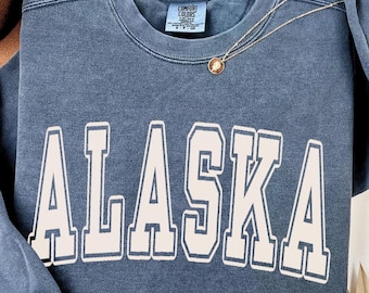Könnte beinhalten: Ein blaues Sweatshirt mit Rundhalsausschnitt und Vintage-Waschung, das das Wort ALASKA in großen, cremefarbenen Blockbuchstaben mit einer dünnen blauen Umrandung zeigt. Das Innenetikett am Kragen zeigt Comfort Colors und Größe S-P-CH.