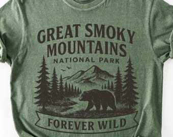 以下が含まれることがあります： セージグリーンの半袖Tシャツで、ダークブラウンのグラフィックプリントが施されています。デザインには「GREAT SMOKY MOUNTAINS NATIONAL PARK」の文字と、松の木、歩くクマ、そして下部に「FOREVER WILD」の文字が描かれた山岳風景が表現されています。