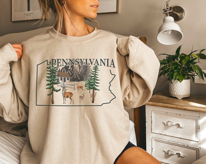 Peut inclure: Un sweat-shirt beige &agrave; col rond arborant le contour vert fonc&eacute; de l'&Eacute;tat de Pennsylvanie. &Agrave; l'int&eacute;rieur du contour, le texte PENNSYLVANIA surmonte un dessin repr&eacute;sentant une cabane, des conif&egrave;res et plusieurs cerfs dans une for&ecirc;t.
