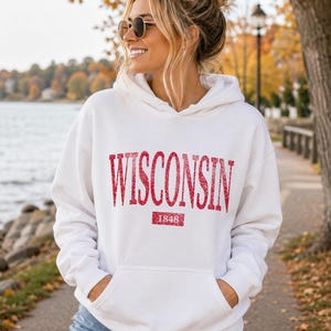 Puede incluir: Una sudadera con capucha blanca que presenta la palabra WISCONSIN en una fuente roja, texturizada y brillante en el pecho. Debajo del texto, un recuadro rojo contiene el a&ntilde;o 1848 en blanco. La sudadera incluye un bolsillo tipo canguro.