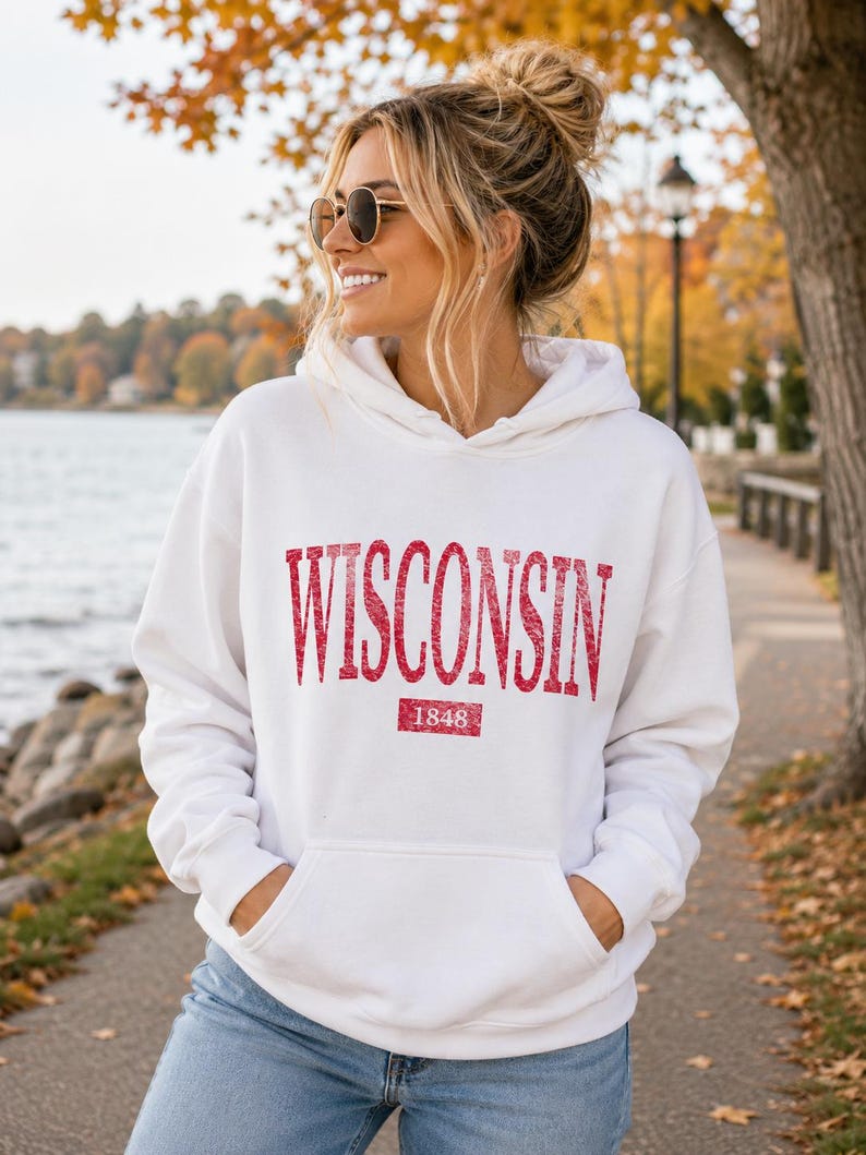 Puede incluir: Una sudadera con capucha blanca que presenta la palabra WISCONSIN en una fuente roja, texturizada y brillante en el pecho. Debajo del texto, un recuadro rojo contiene el a&ntilde;o 1848 en blanco. La sudadera incluye un bolsillo tipo canguro.