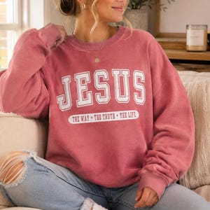 May include: A dusty rose crewneck sweatshirt featuring the white text JESUS in a collegiate font, with the phrase THE WAY THE TRUTH THE LIFE printed in white below it. The garment has long sleeves and a relaxed fit, paired with light blue denim jeans.