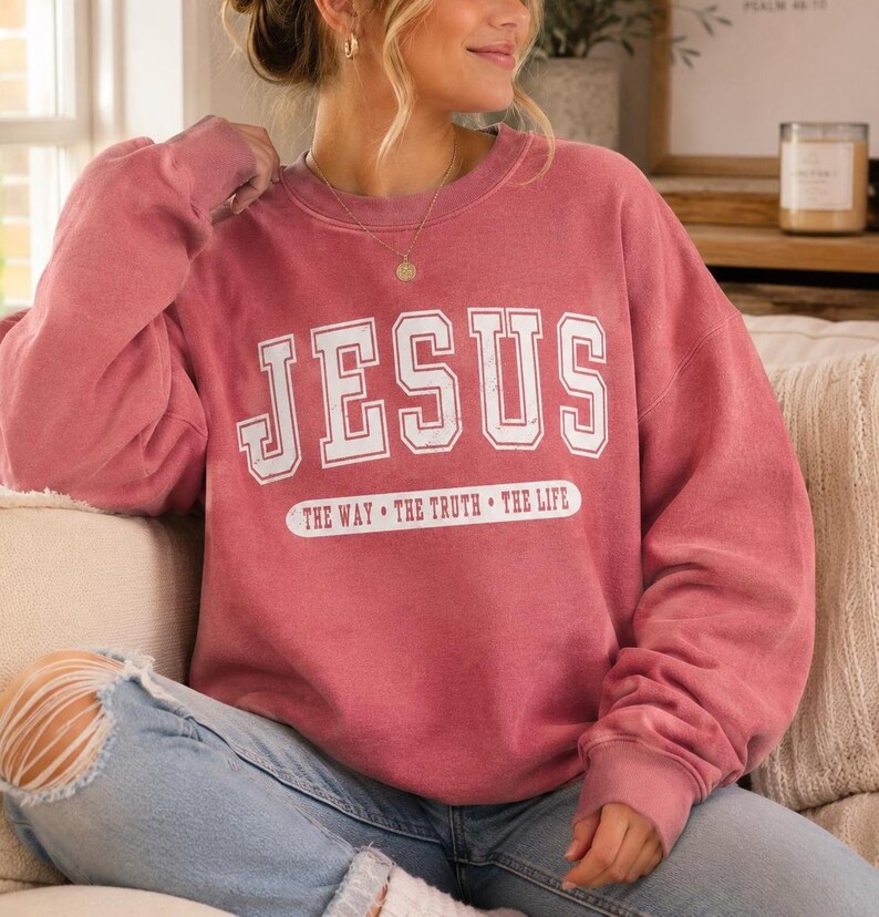 May include: A dusty rose crewneck sweatshirt featuring the white text JESUS in a collegiate font, with the phrase THE WAY THE TRUTH THE LIFE printed in white below it. The garment has long sleeves and a relaxed fit, paired with light blue denim jeans.