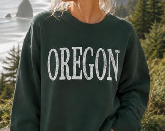 Può includere: Una felpa girocollo verde bosco scuro con la parola OREGON stampata sul petto in un grande carattere serif bianco dall'aspetto vissuto. Il capo presenta maniche lunghe e bordi a costine su scollo, polsini e orlo inferiore.