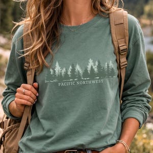 May include: A sage green long-sleeve crewneck shirt featuring a white graphic print of a coniferous forest silhouette above the text PACIFIC NORTHWEST. The shirt is worn with a tan backpack, showcasing a casual outdoor-inspired aesthetic.