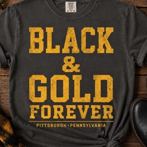 以下が含まれることがあります： BLACK & GOLD FOREVERという掠れた黄色のブロック体文字と、その下にPITTSBURGH • PENNSYLVANIAという文字がプリントされたチャコールグレーの半袖Tシャツ。クルーネックで、襟元にはComfort Colorsのブランドラベルが付いています。