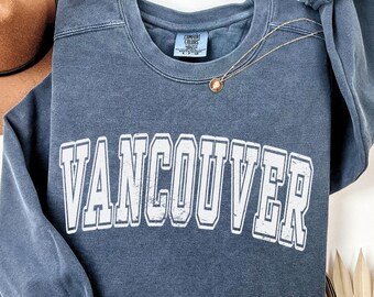May include: A faded navy blue sweatshirt with the word "VANCOUVER" in large, white, block letters. The sweatshirt has a crew neck and long sleeves. A gold necklace with a pendant is draped across the front.