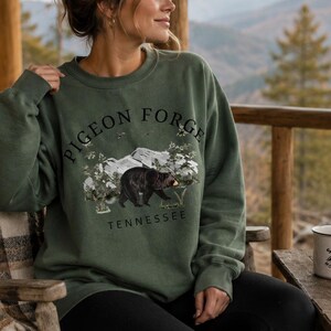 K&ouml;nnte beinhalten: Ein olivgr&uuml;nes Sweatshirt mit Rundhalsausschnitt und einem schwarzen Grafikdruck eines Schwarzb&auml;ren, der durch einen Wald mit Bergen im Hintergrund l&auml;uft. Der Text PIGEON FORGE steht in einem Bogen &uuml;ber dem B&auml;ren und TENNESSEE darunter gedruckt.