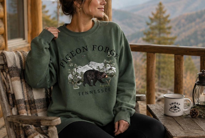 K&ouml;nnte beinhalten: Ein olivgr&uuml;nes Sweatshirt mit Rundhalsausschnitt und einem schwarzen Grafikdruck eines Schwarzb&auml;ren, der durch einen Wald mit Bergen im Hintergrund l&auml;uft. Der Text PIGEON FORGE steht in einem Bogen &uuml;ber dem B&auml;ren und TENNESSEE darunter gedruckt.