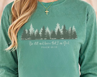 May include: A long-sleeve crewneck sweatshirt in a muted green shade. The front features a graphic of a forest with pine trees in varying shades of green and gray. Below the trees, the text reads: Be still and know that I am God PSALM 46:10.