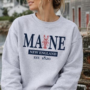 May include: A light gray crewneck sweatshirt featuring the dark blue text MAINE with a red lobster graphic replacing the letter I. Below, the text reads NEW ENGLAND in a dark blue box, followed by the text EST. 1820 in dark blue lettering.
