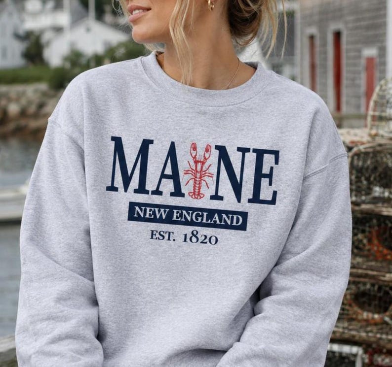 May include: A light gray crewneck sweatshirt featuring the dark blue text MAINE with a red lobster graphic replacing the letter I. Below, the text reads NEW ENGLAND in a dark blue box, followed by the text EST. 1820 in dark blue lettering.