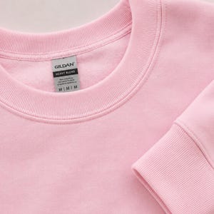 Pode incluir: Uma camisola de gola redonda em rosa claro com gola e punhos canelados. Uma etiqueta cinzenta Gildan Heavy Blend est&aacute; cosida no interior da gola, com o texto Gildan, Heavy Blend e o tamanho M impressos a preto na etiqueta.