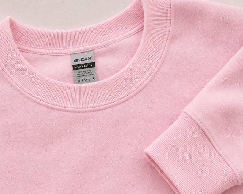 Pode incluir: Uma camisola de gola redonda em rosa claro com gola e punhos canelados. Uma etiqueta cinzenta Gildan Heavy Blend est&aacute; cosida no interior da gola, com o texto Gildan, Heavy Blend e o tamanho M impressos a preto na etiqueta.