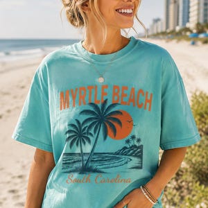 May include: A turquoise short-sleeve t-shirt featuring a graphic print of palm trees, an orange sun, and the text Myrtle Beach South Carolina in orange lettering. The shirt has a crew neckline and a relaxed fit, showcasing a coastal beach scene.